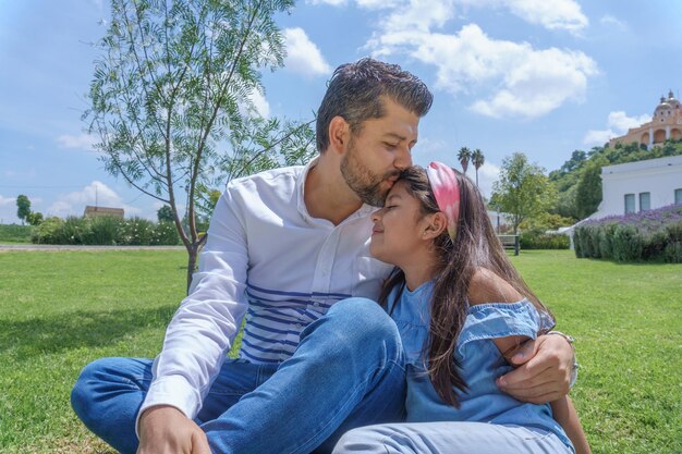 公園の緑の芝生に座って娘の頭にキスをする父親