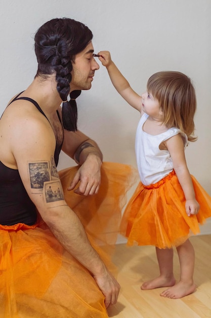 Father and little girl are wearing matching ballerina dresses. girl applying make-up to her father