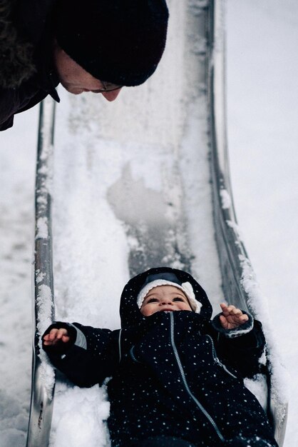 雪で覆われたスライドで遊んでいる赤ちゃんの女の子を見ている父親