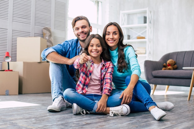 Отец, мать и дочь обнимаются, сидя на полу своей недавно купленной квартиры, держа ключи и широко улыбаясь.