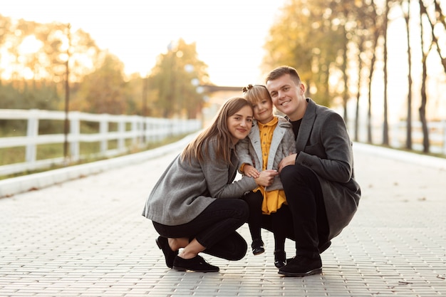 Father and mother are holding little daughter by the hands and walking in the autumn park