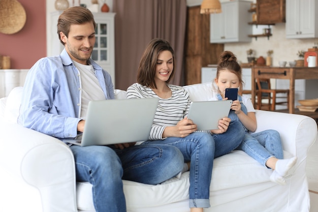 Father, mother and daughter using electronic devices sitting on sofa at living room.