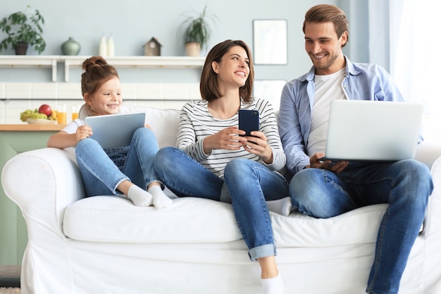 Padre, madre e figlia che utilizzano dispositivi elettronici seduti sul divano in soggiorno.