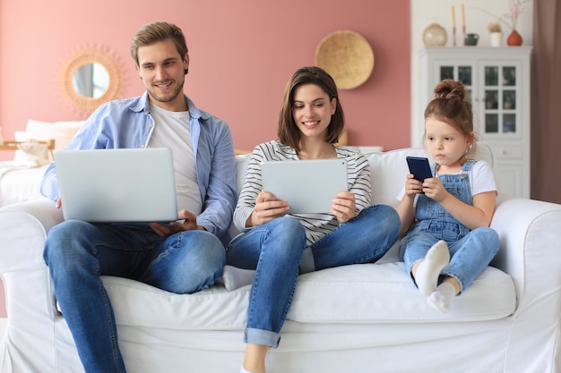 Father, mother and daughter using electronic devices sitting on sofa at living room.