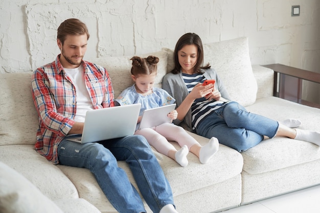 Padre, madre e figlia che utilizzano dispositivi elettronici seduti sul divano in soggiorno.