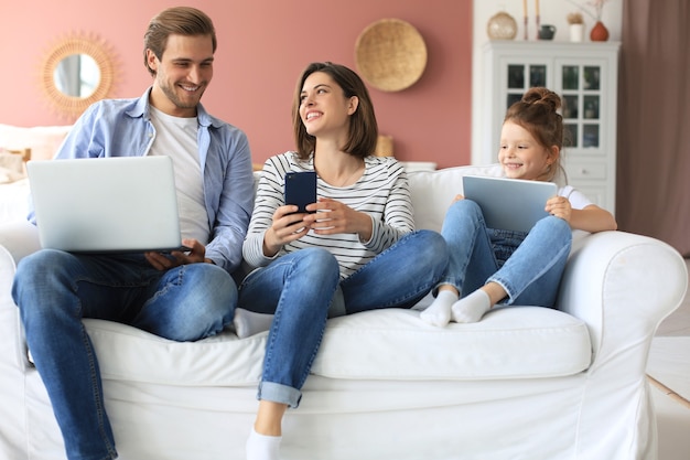 Padre, madre e figlia che utilizzano dispositivi elettronici seduti sul divano in soggiorno.