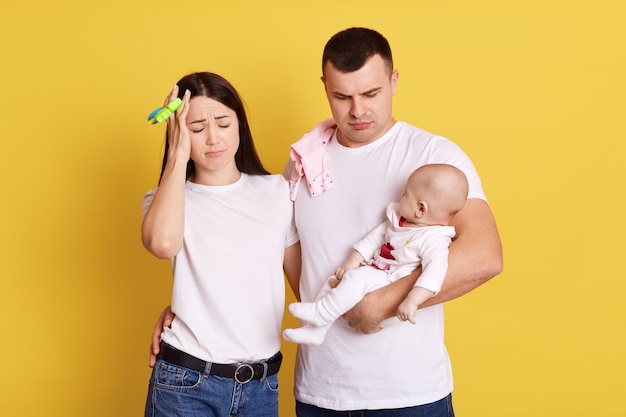 Father, mother and their infant child standing against yellow wall. Busy tired dad and mom poses with baby on hands, hesitant mommy stands near and keeps hand on forehead. Parents care about newborn.