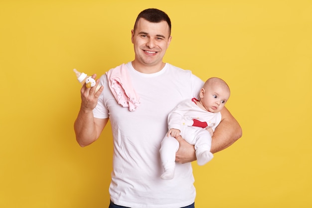 Father needs feeding baby, wearing white casual t shirt, holding newborn daughter and feed bottle in hands, being happy, handsome male on paternity leave.