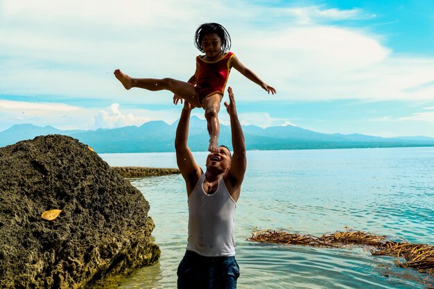Photo father playing with daughter while standing at beach against sky