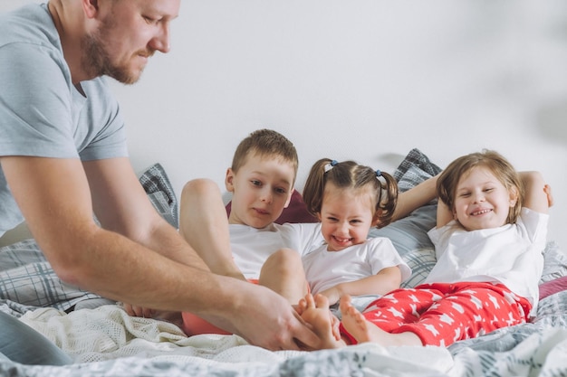 Отец играет с тремя детьми на кровати. Папа щекочет детям ножки. Семья папы, двух девочек и мальчика