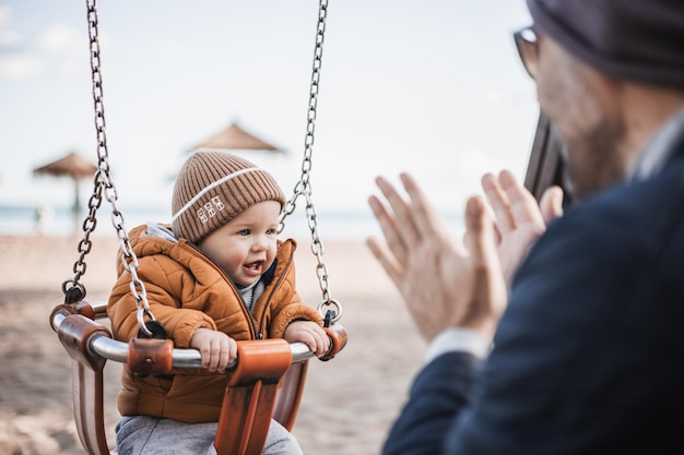 Father pushing hir cheerful infant baby boy child on a swing on sandy beach playground outdoors on nice sunny cold winter day in Malaga Spain