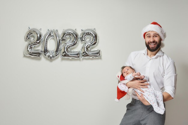 A father in a red velvet Santa hat is holding his infant daughter near silver balloons in the shape of 2022. A bearded dad with his little child is smiling at a New year party.