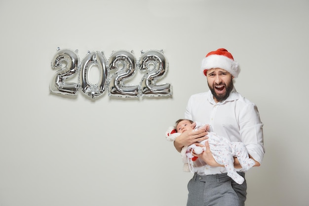 A father in a red velvet Santa hat is holding his infant daughter near silver balloons in the shape of 2022. A dad is hugging his little child and screaming at a New year party.