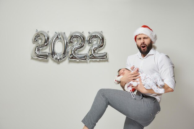 A father in a red velvet Santa hat is holding his infant daughter near silver balloons in the shape of 2022. A dad with his little child is lifting his leg at a New year party.