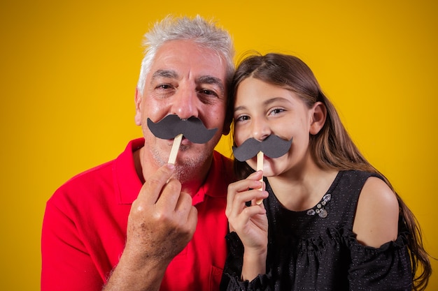 Foto festa del papà. figlia felice che tiene un po' di baffi di carta con suo padre su sfondo giallo. figlia e papà con i baffi in vacanza