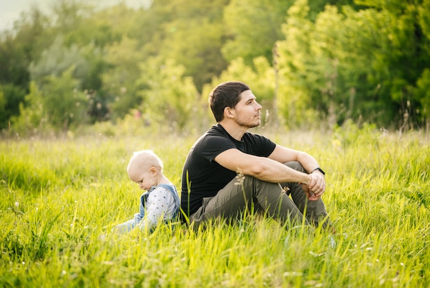 Father's day. A man and his daughter enjoy a vacation in a green park in the evening at sunset in summer, sitting back to back in the grass in a meadow.