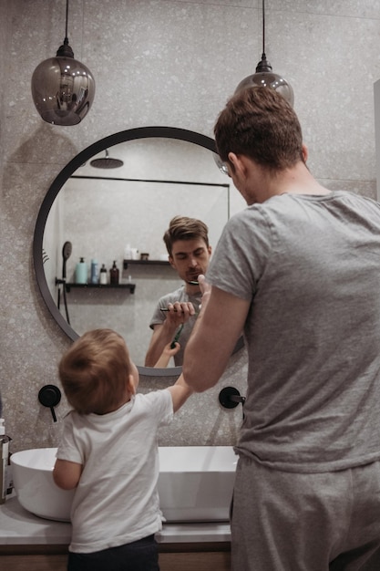 A father and son brushing their teeth together in a bathroom together while looking at the reflectio