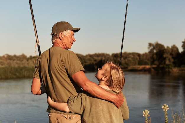 Father and son go fishing place, standing near lake and hugging, looking at each other, wearing green clothing, family spending time together in open air and enjoying beautiful nature.