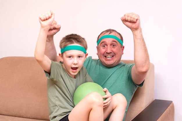 A father and son in green bandanas and t shirts sit on a beige sofa and cheer emotionally for the football team on TV