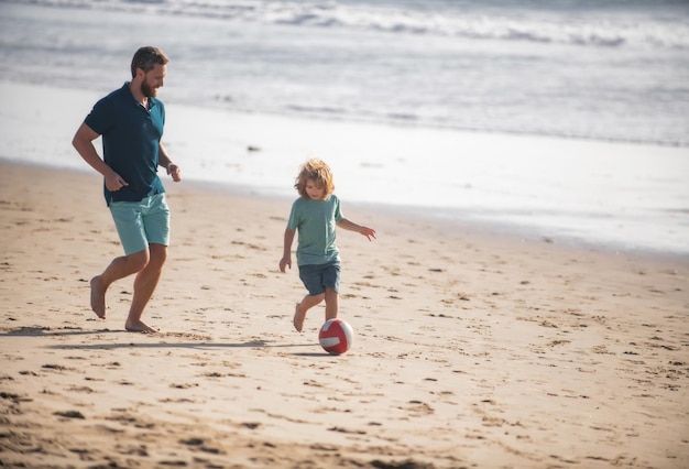 Father and son play soccer or football on the beach on summer family holidays dad and child enjoying