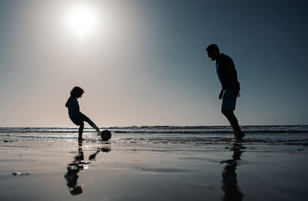 Father and son play soccer or football on the beach on summer family holidays