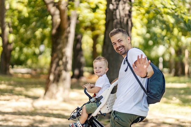 A father and son ride a bicycle and wave at the camera. Toddlers sit in a basket with bicycles and wander around the park by bicycle. Summer sunny family day, time of family connection. Active family
