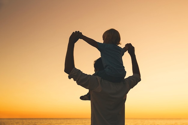 Father and son silhouettes at sunset sky Loving family and summer vacation Man and kid boy playing together outdoors on a sea beach Dad carrying child on his back with raised arms up on nature