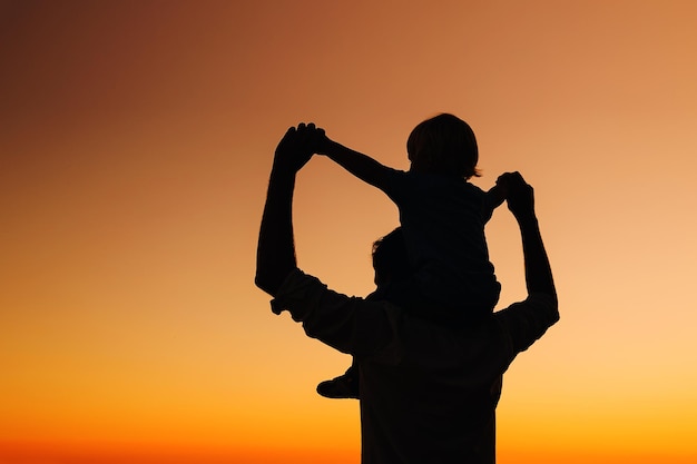 Photo father and son silhouettes at sunset sky loving family and summer vacation man and kid boy playing together outdoors on a sea beach dad carrying child on his back with raised arms up on nature