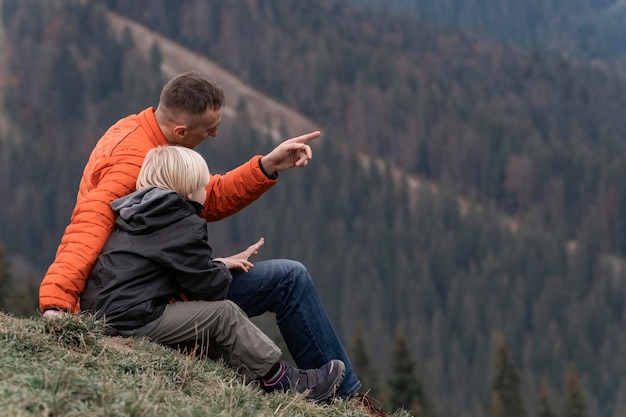 아버지와 아들은 숲 배경의 언덕에 앉아 있습니다 아이들과 함께 트레킹 및 하이킹 산에서 주말 하이킹
