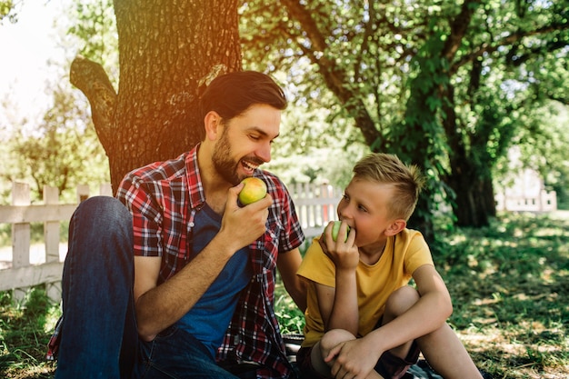 father and son sitting together under tree and eating apple