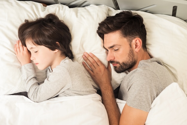 Father and son sleep together on the bed in their house.