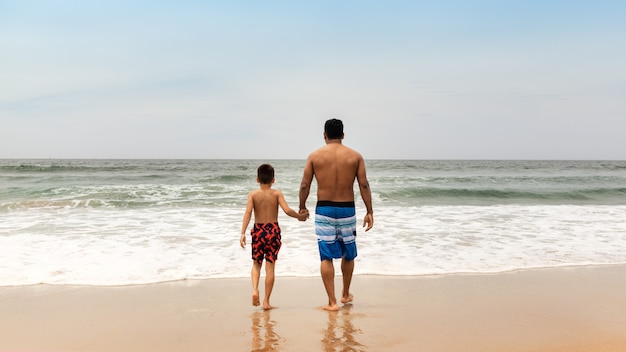 Padre e figlio in piedi sulla spiaggia tenendosi per mano e guardando il mare