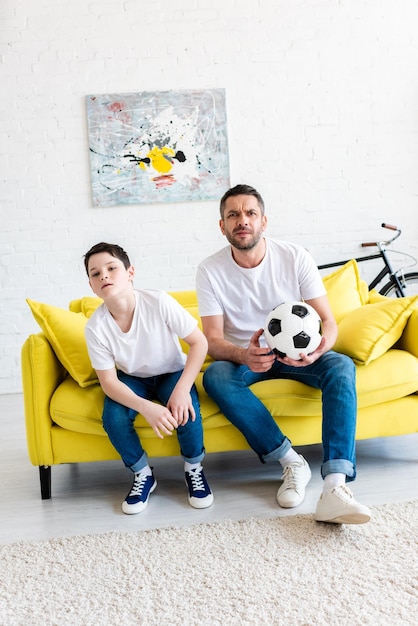 Father and son watching sports match on couch with soccer ball at home