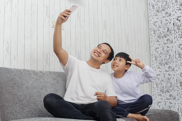 Father and son wearing white t shirt taking photo togehther with phone on the sofa suitable for app