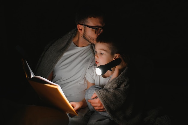 Father and son with flashlight reading book under blanket at home