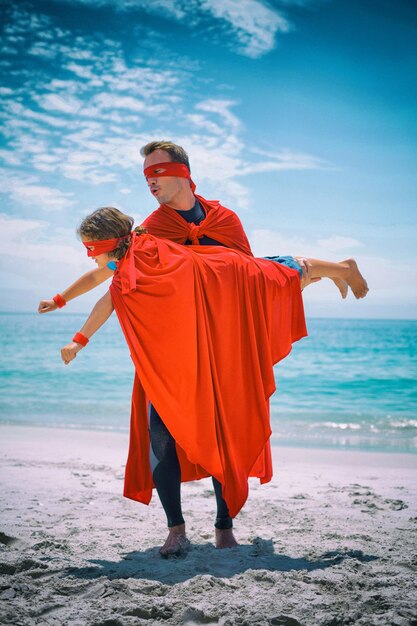 Father in superhero costume lifting son at sea shore