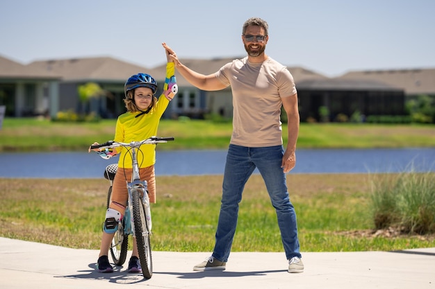 父は息子をサポートします。スポーティな家族の父と息子が公園で自転車に乗って、安全ヘルメットに乗って子供をサポートします。