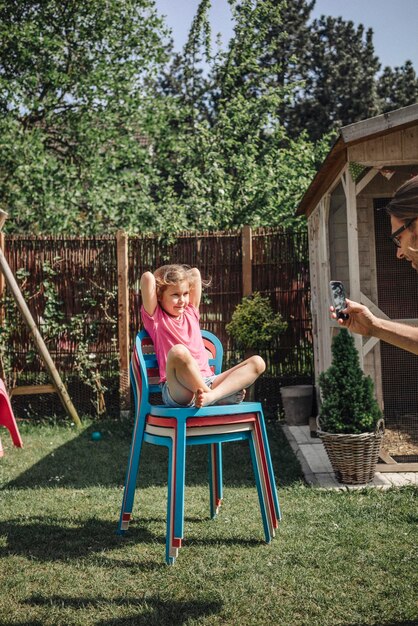 Father taking cell phone picture of daughter sitting on chair in garden