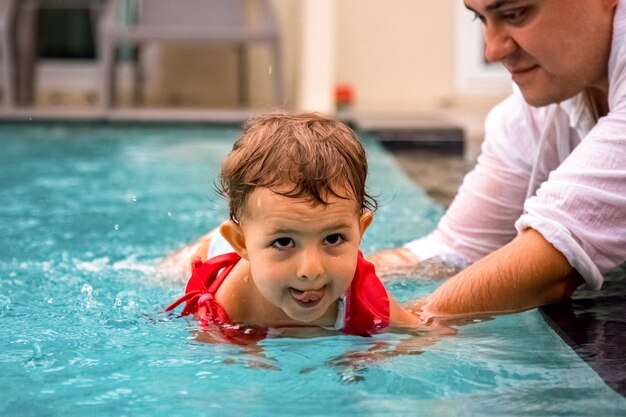 father teaches cute toddler little girl in swimsuit to swim in the pool holding his hand.