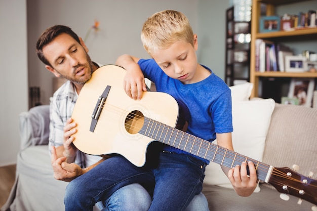 Father teaching son to play guitar while sitting on sofa