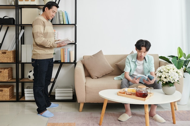 Photo father and teenage son reading books and drinking tea with snacks when staying home due to covid-19 pandemic