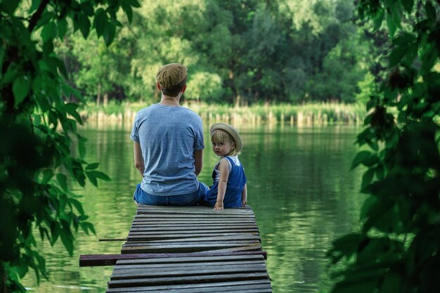 Father and a tiny girl in dungarees are sitting on the bridge and looking at the camera