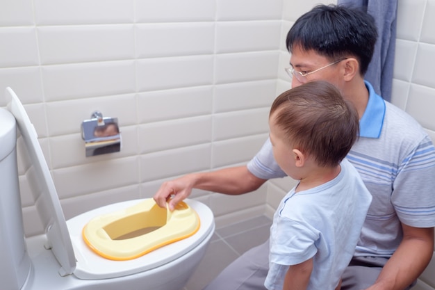 Photo father training sleepy son to use toilet in bathroom, asian toddler boy sitting on toilet with kid bathroom accessory