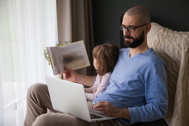 Father trying to work on his laptop sitting at home with his little daughter, while she's reading a book