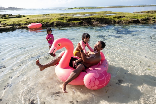 Father and two daughters ride flamingo buoy on the beach