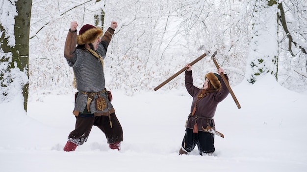 Foto padre vichingo con suo figlio nella foresta invernale. il padre insegna a suo figlio a combattere, tenendo in mano l'ascia. si vestivano con abiti medievali.