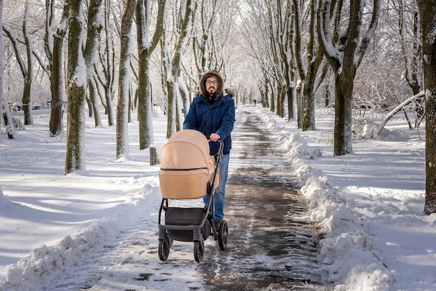 雪に覆われた公園でベージュのベビーカーと一緒に歩いている父寒い美しい冬の日のレクリエーション