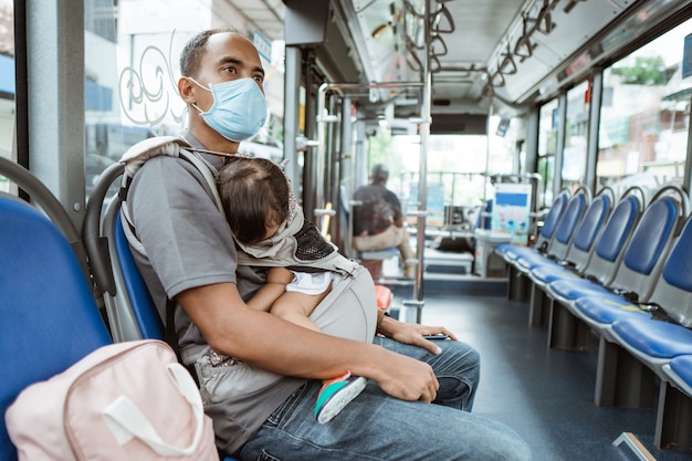 Un padre che indossa una maschera siede su una panchina con in braccio una piccola bambina che dorme sull'autobus lungo la strada