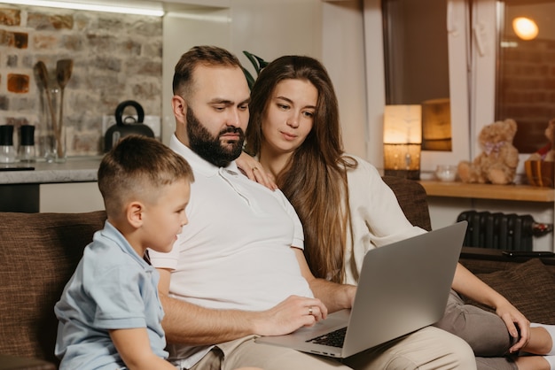 Бородатый отец удаленно работает на ноутбуке, а его сын и жена смотрят на него. Семья вечером на диване. Папа работает онлайн на компьютере между родственниками дома