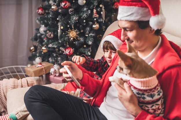 Father with child kid son and puppy dog in santa hats at christmas holidays sitting on a couch in